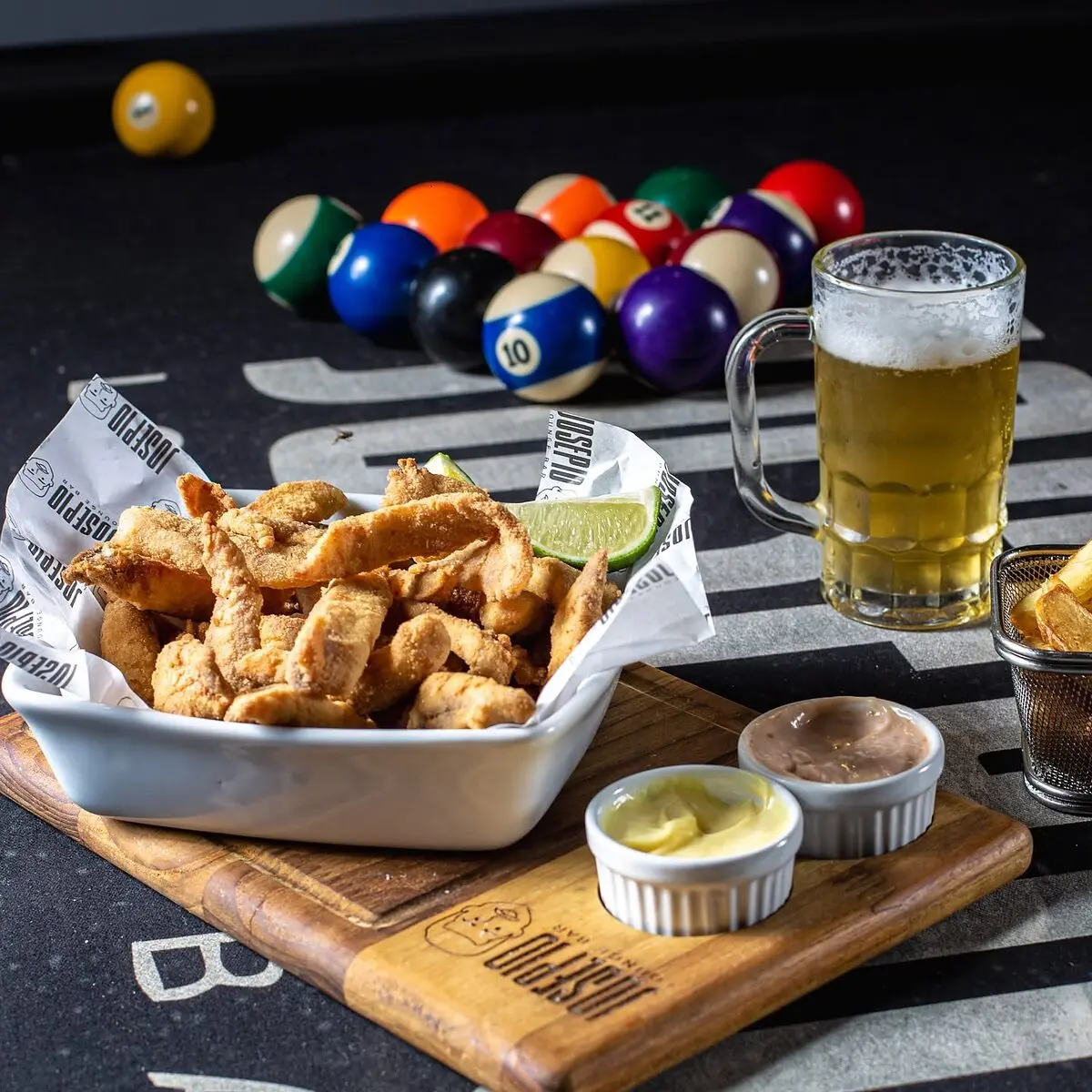 Isca de peixe com limão e molhos, ao lado de um chopp na mesa de sinuca